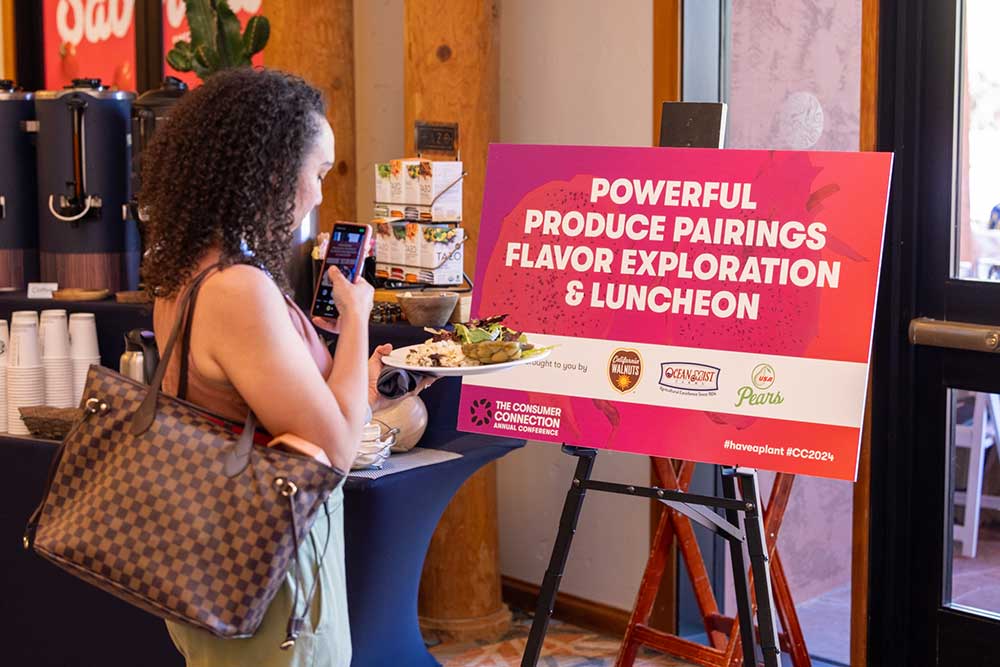 A woman holding a plate of food takes a photo of a sign that reads: Powerful Produce Pairings Flavor Exploration & Luncheon.